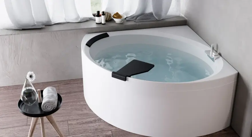 Una vasca da bagno angolare bianca, piena d'acqua, con accessori da bagno e un fiore su un tavolino. Un'esperienza novellini divina.