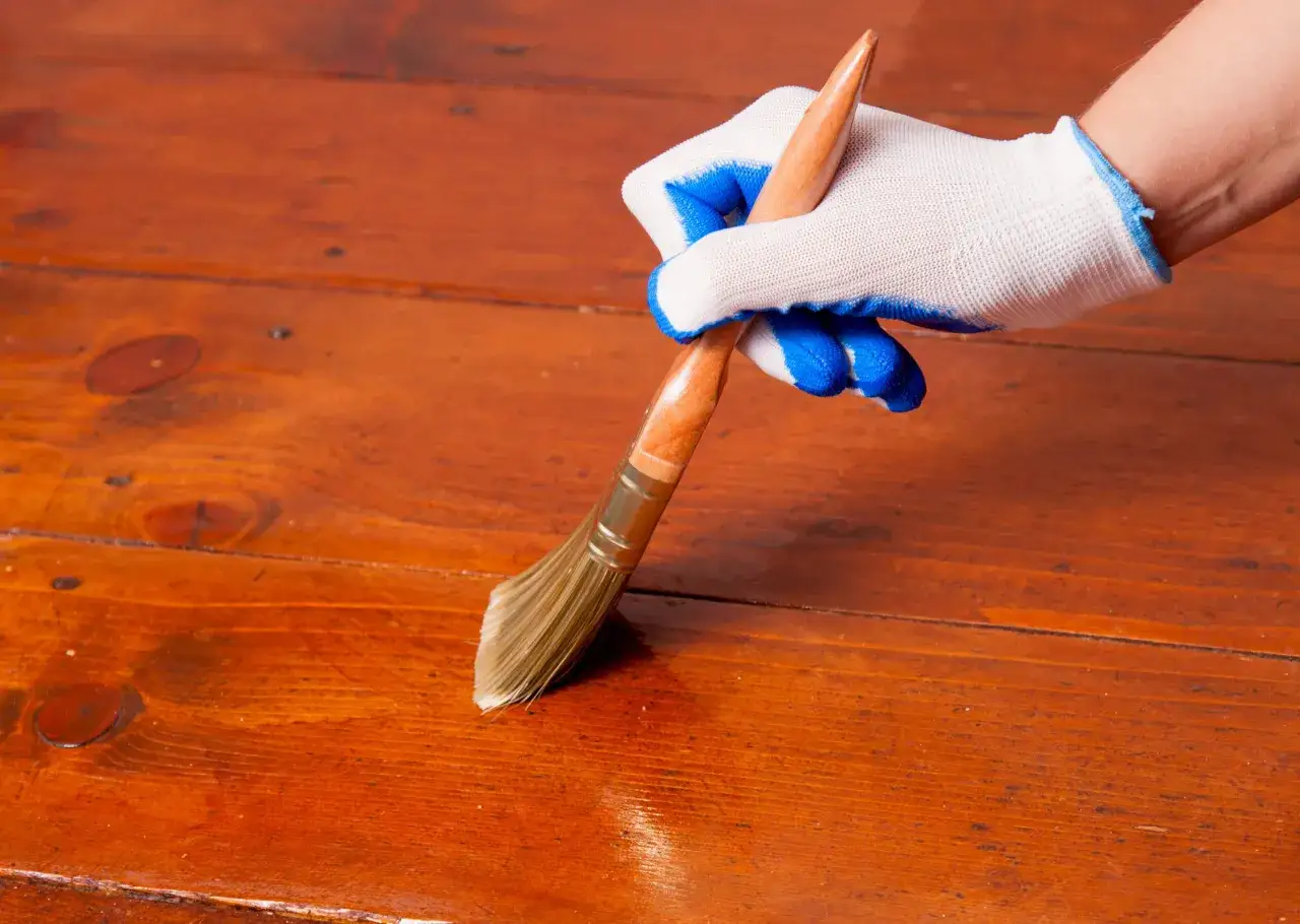 Mano con guanto bianco e blu applica vernice trasparente su un parquet in legno scuro.