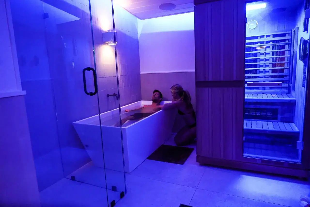Coppia in un bagno moderno con vasca e sauna. Lei accarezza lui nella vasca, un momento di relax che ricorda la doccia svedese.