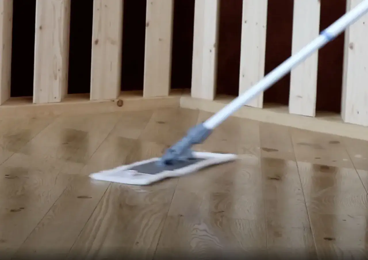 Come togliere la cera dal parquet verniciato: un mocio pulisce il pavimento in legno lucido vicino a una ringhiera in legno.