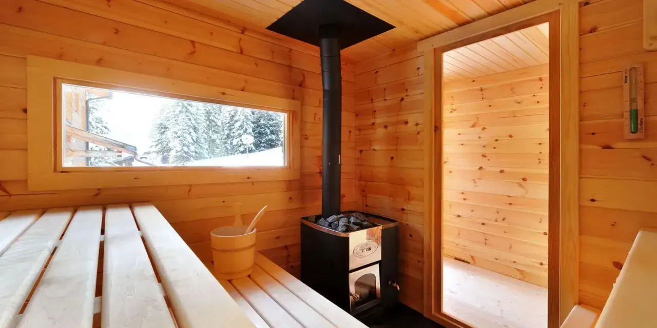 Interno di una sauna benessere in legno con stufa, secchiello e finestra che mostra un paesaggio innevato.