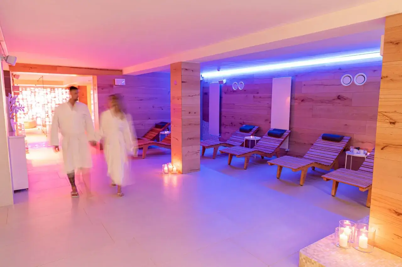 Coppia in accappatoio cammina in una spa illuminata da luci soffuse. Lettini prendisole e candele creano un'atmosfera rilassante, perfetta per scoprire cosa si fa in un centro benessere.