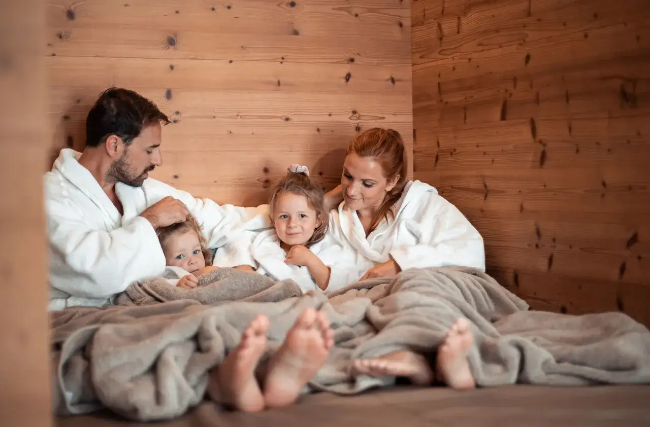 Famiglia in accappatoio, con bambini, in una sauna di legno. Non c'&egrave; un'et&agrave; minima per godersi il relax in sauna.