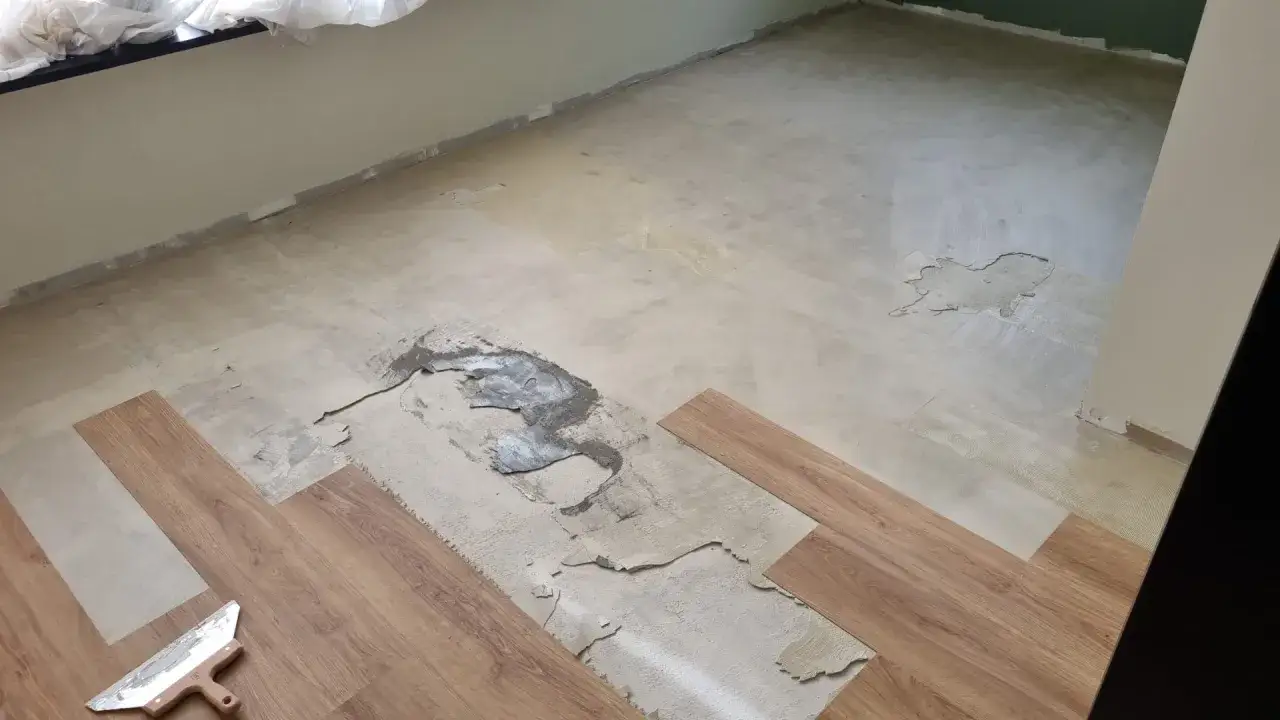 Il pavimento in PVC si solleva in alcune zone, mostrando il sottofondo danneggiato. Si vedono nuove doghe in legno e una spatola.