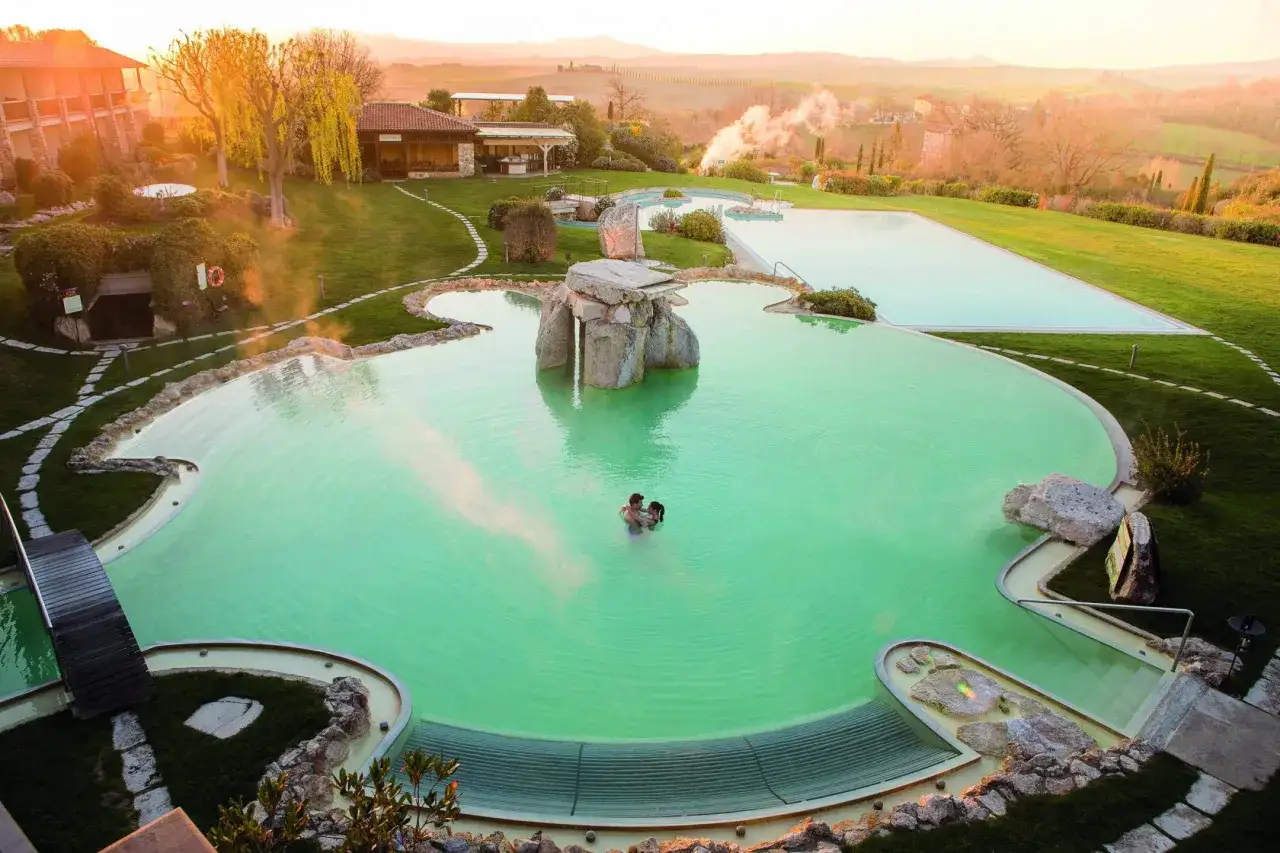 Coppia abbracciata in una piscina termale verde smeraldo, con un paesaggio collinare al tramonto. La differenza tra spa e terme &egrave; chiara: qui si vive un'esperienza rigenerante.
