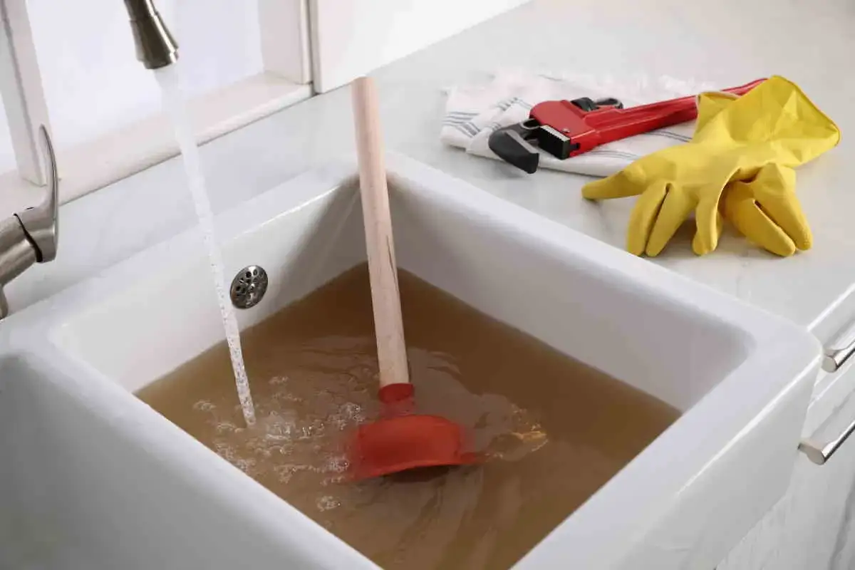 Vasca da bagno piena d'acqua torbida con uno sturalavandini pronto all'uso. Chiave inglese e guanti gialli sul bordo, per risolvere come sturare la vasca da bagno.