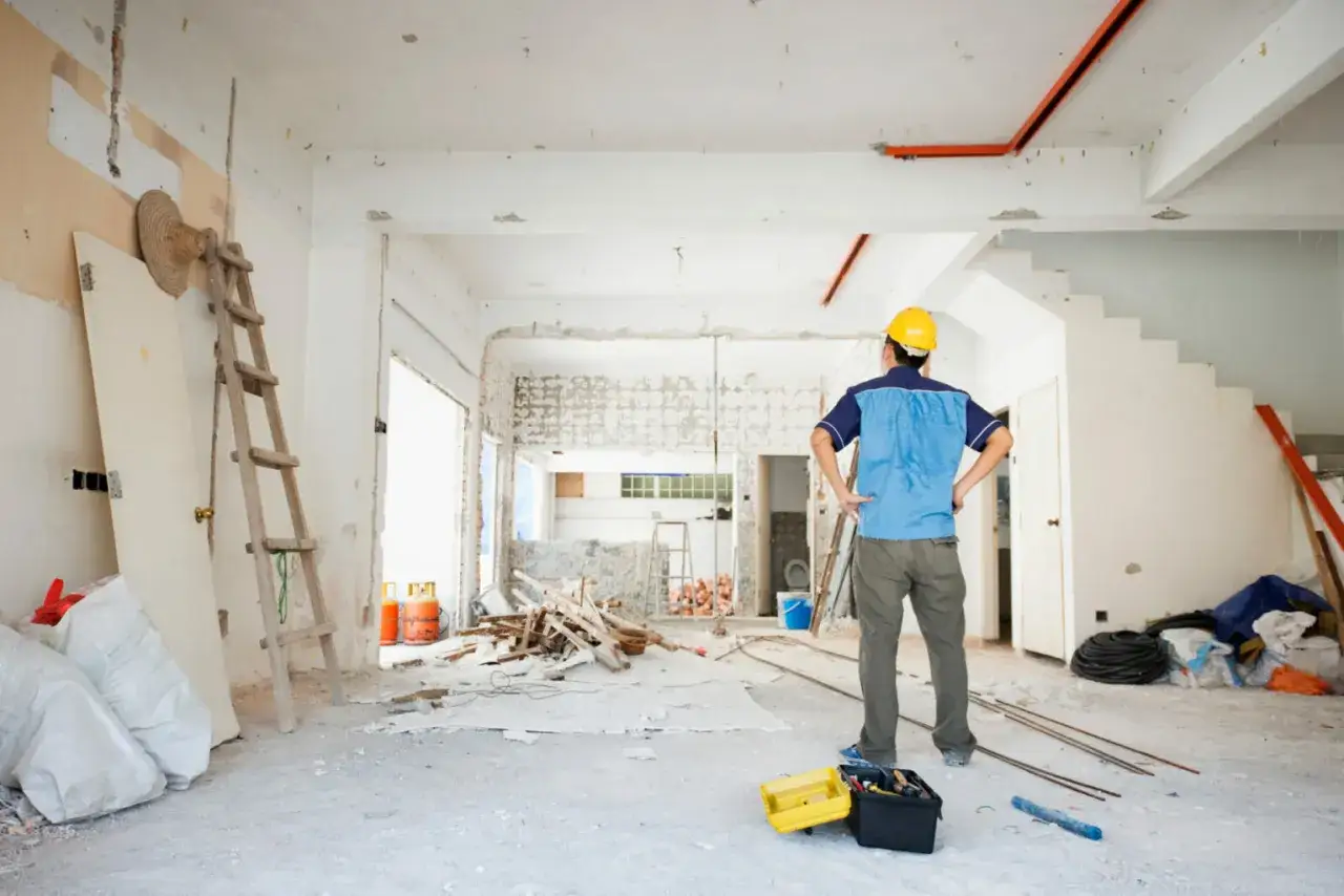 Operaio con casco giallo osserva il cantiere, pronto a rimuovere pavimento sovrapposto. Scale, sacchi e detriti indicano lavori in corso.