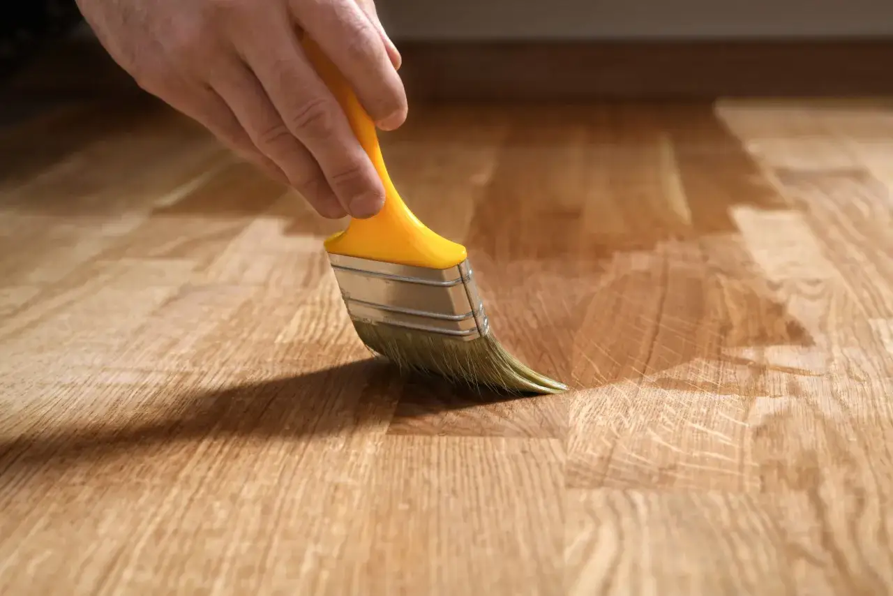 Mano con pennello giallo applica un liquido trasparente per pulire parquet con alcool, evidenziando le venature del legno.