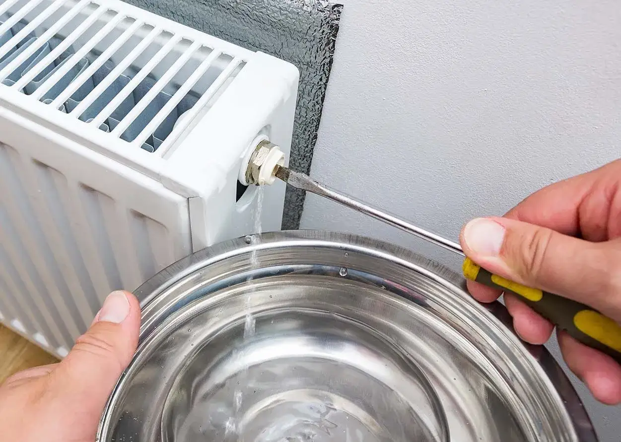 Un rubinetto del termosifone perde acqua in una bacinella mentre qualcuno cerca di stringerlo con un cacciavite.