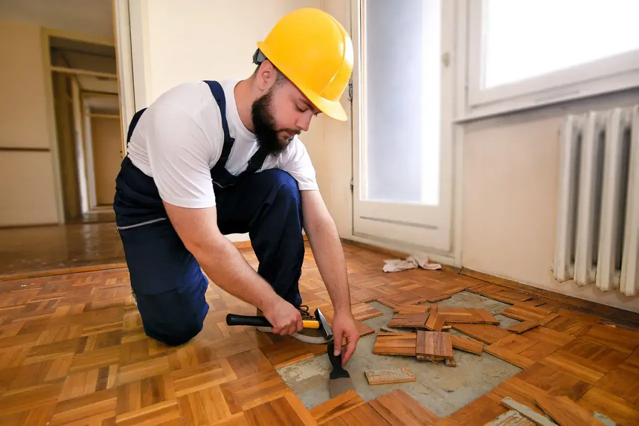Operaio con casco giallo impegnato a rimuovere parquet incollato con scalpello e martello.